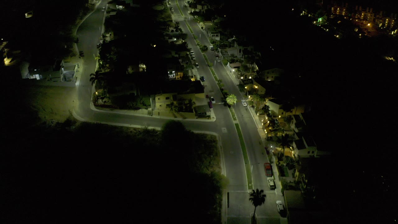 Early frame of a Los Cabos, Mexico night aerial showing a lit road curving into an intersection, nearby homes, parked cars, trees, and no visible people.