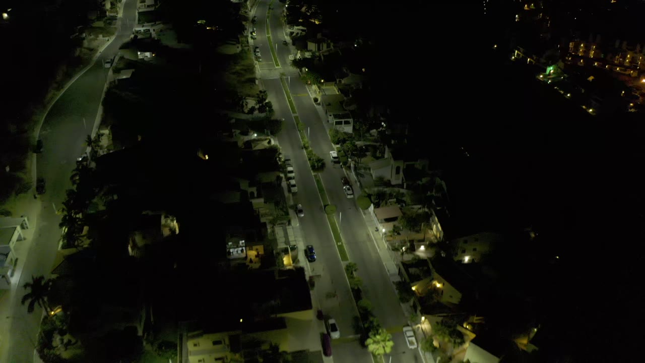 Night drone-like view in Los Cabos, Mexico, with the road slightly shifted in frame, streetlights on pavement, houses, trees, parked cars, and no visible people.