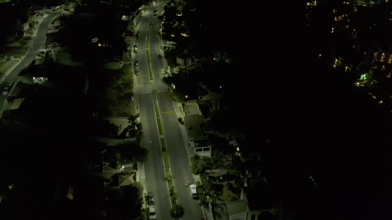 A later aerial frame shows the Los Cabos road continuing through darkness, with illuminated pavement, sidewalks, parked vehicles, tree shadows, and no visible people.