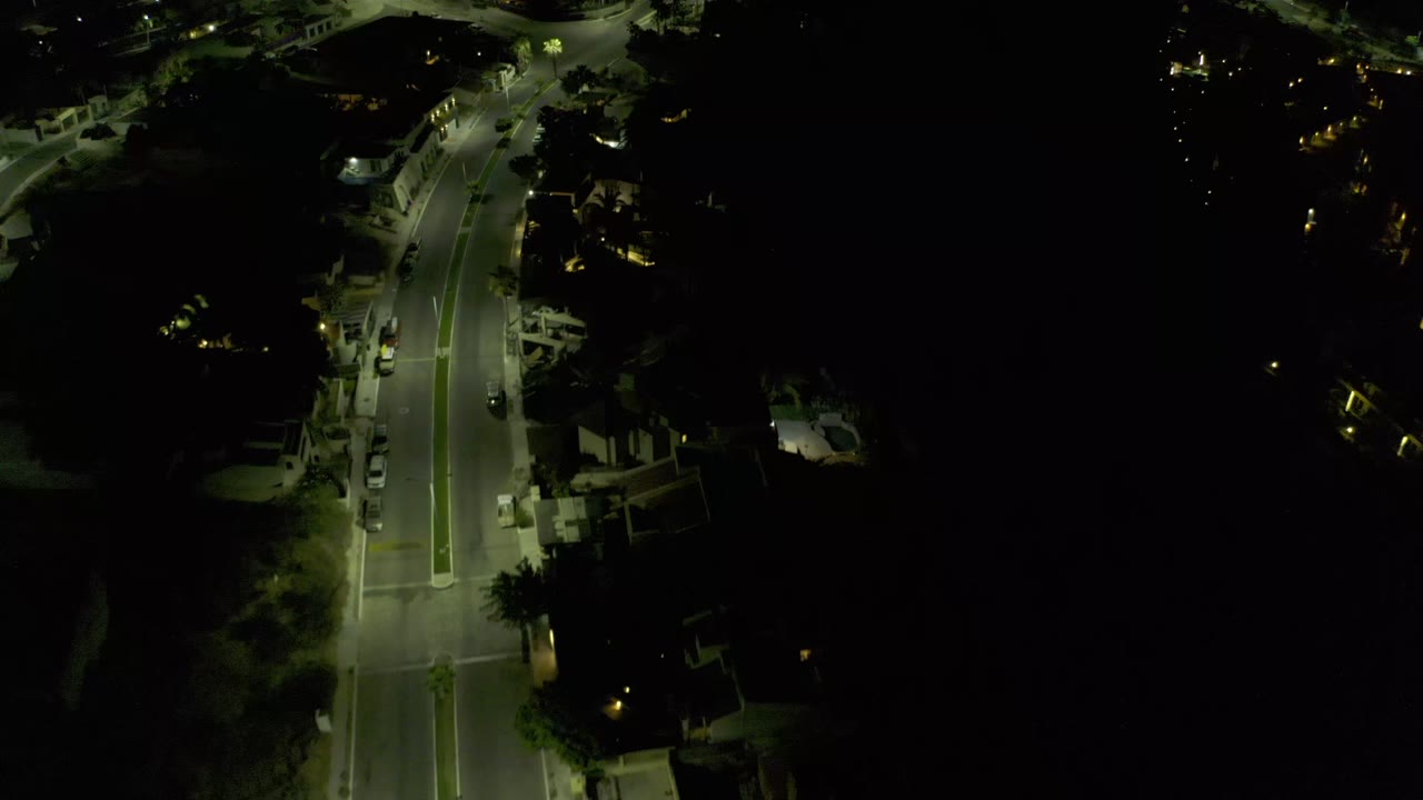Bird's-eye nighttime frame over a Los Cabos road segment, with greenish streetlight pools, a slight bend near the top, parked vehicles, and no visible people.