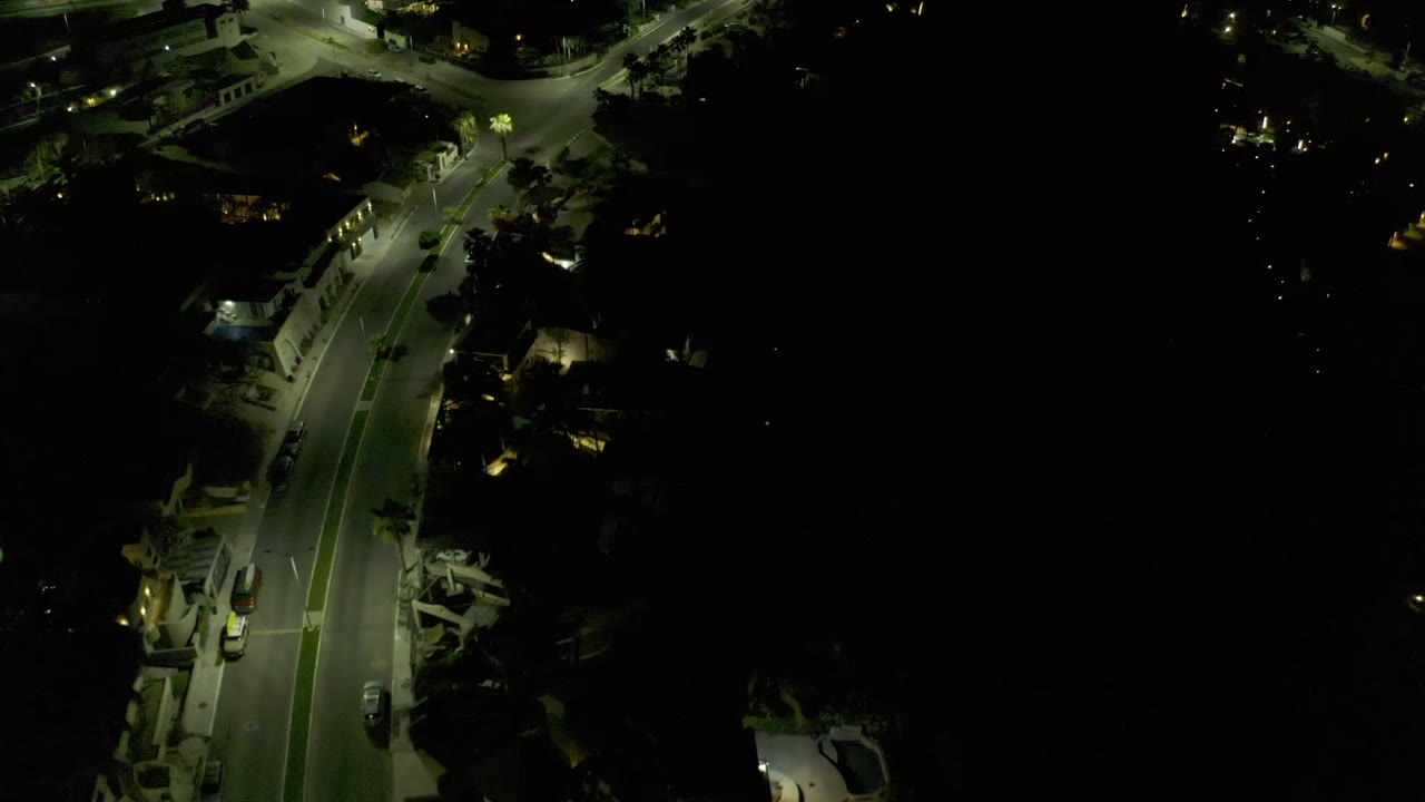 High night view over Los Cabos showing the same road bend, bright streetlight pools, shadowed homes, roadside trees, vehicles, and no visible people.