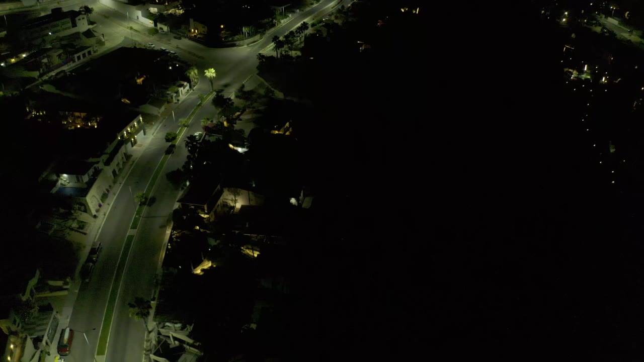 High night view over a Los Cabos roadway, with the lit pavement bending through dark surrounding buildings and no visible people.