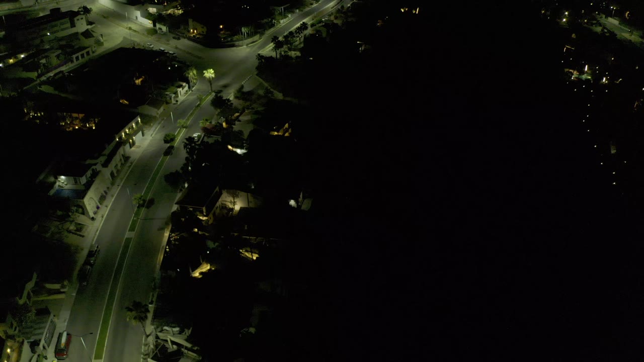 Aerial nighttime frame in Los Cabos showing a quiet lit street, palm trees near the curb, and mostly black surroundings with no visible people.