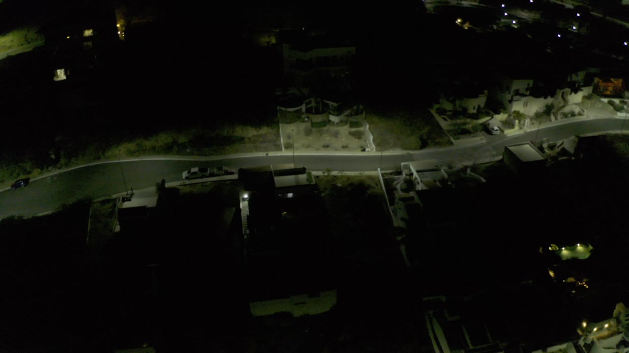High night aerial over Los Cabos, Mexico, showing a curved residential road, walled buildings, scattered lights, and no visible people.