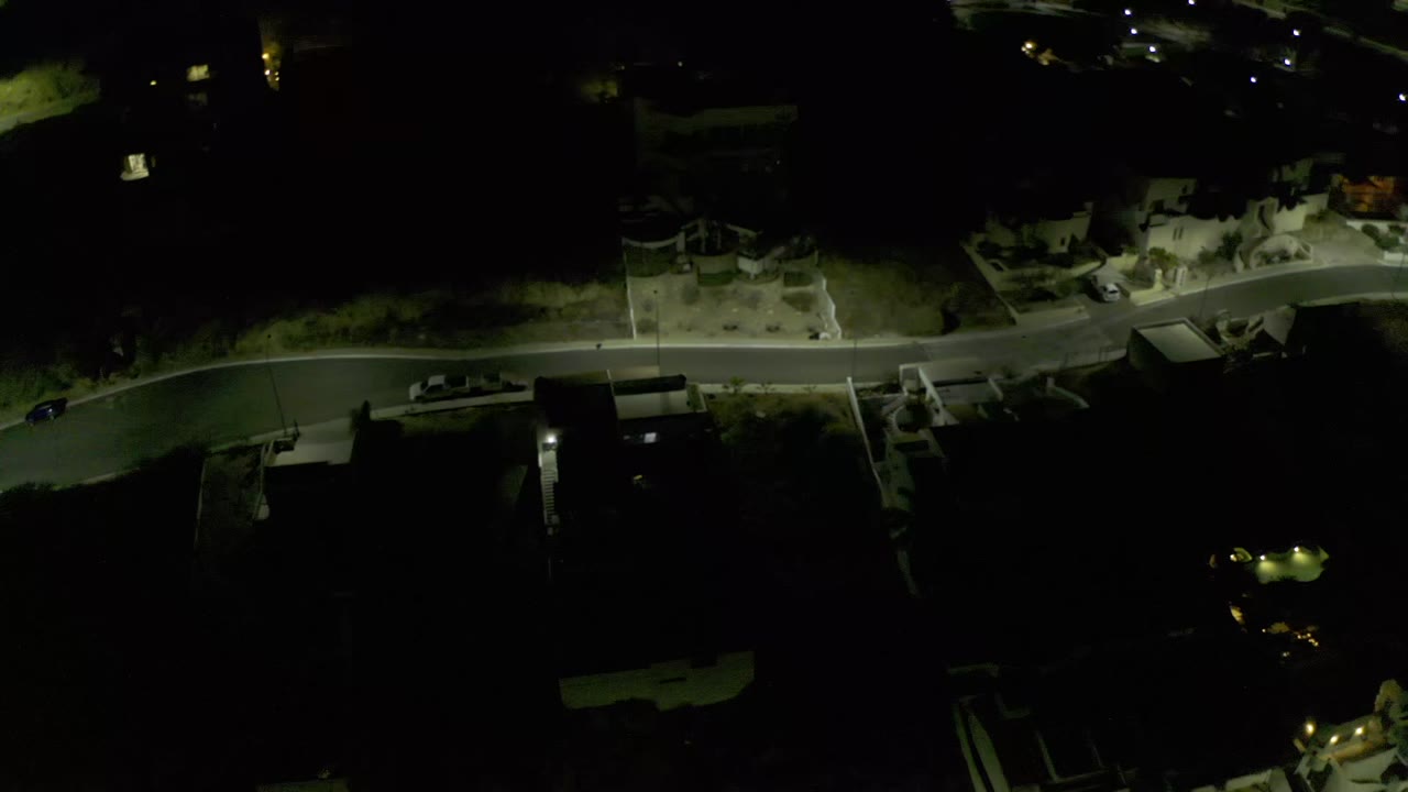 Drone view over Los Cabos, Mexico, with the road crossing the frame, pale building walls near the street, deep shadowed lots, and no visible people.