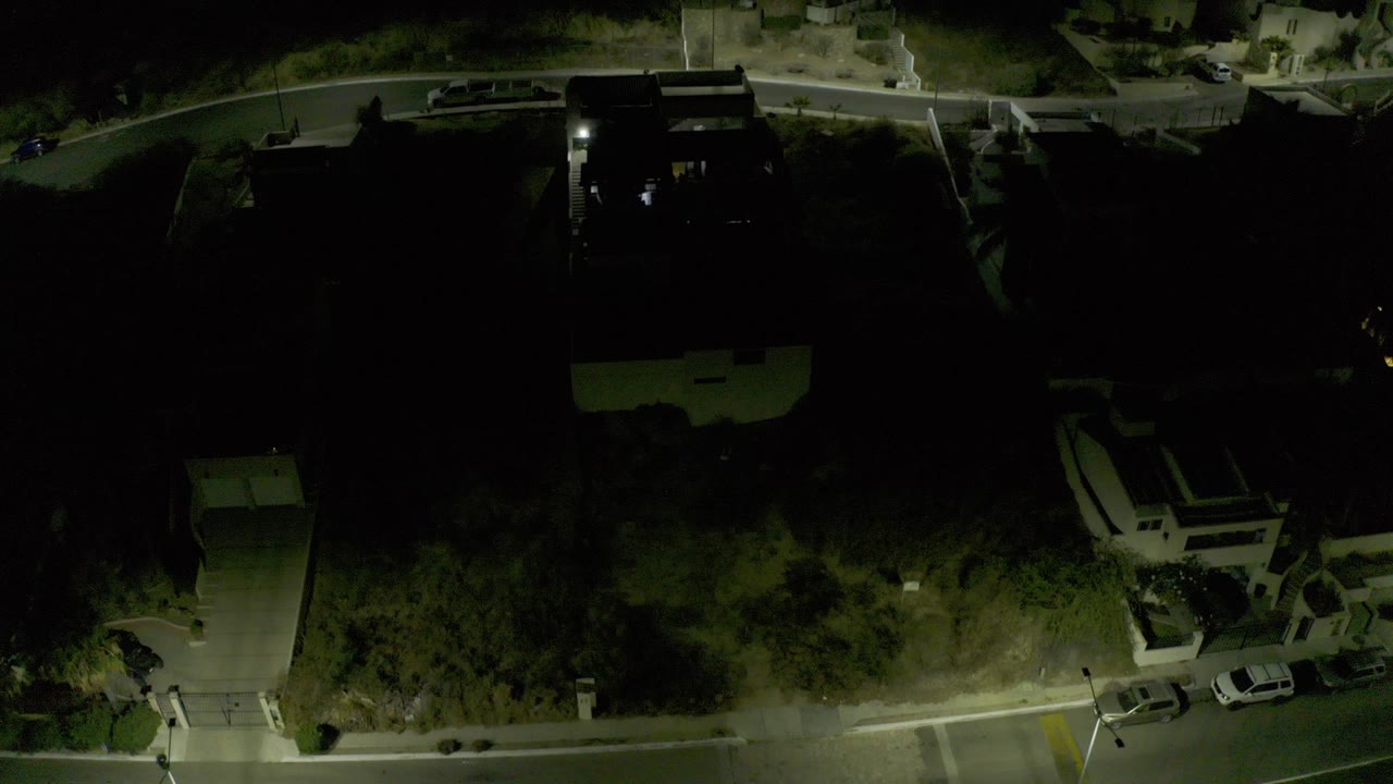 Dark overhead drone view in Los Cabos, Mexico, showing a residential street, houses, parked cars, trees, and no visible people at the opening moment.