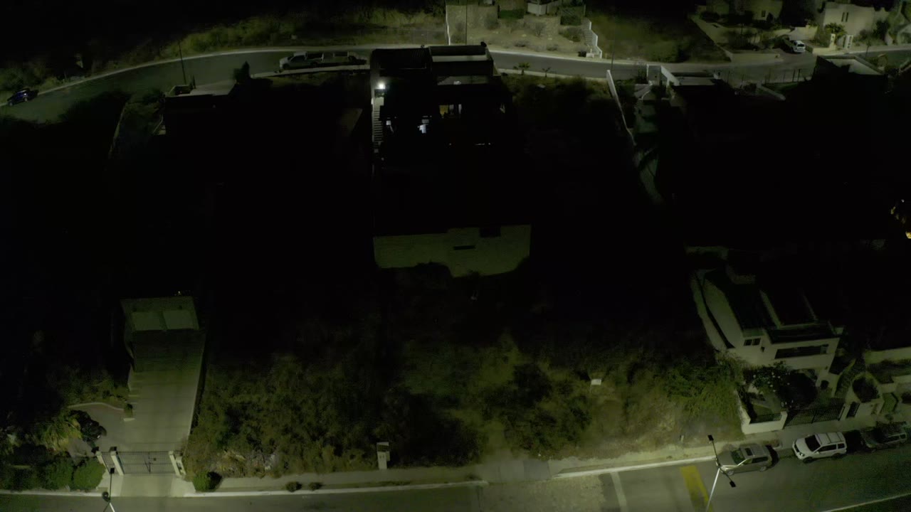Night aerial frame in Los Cabos, Mexico, with a dark residential block, visible driveways, parked cars near the curb, trees, and no visible people.