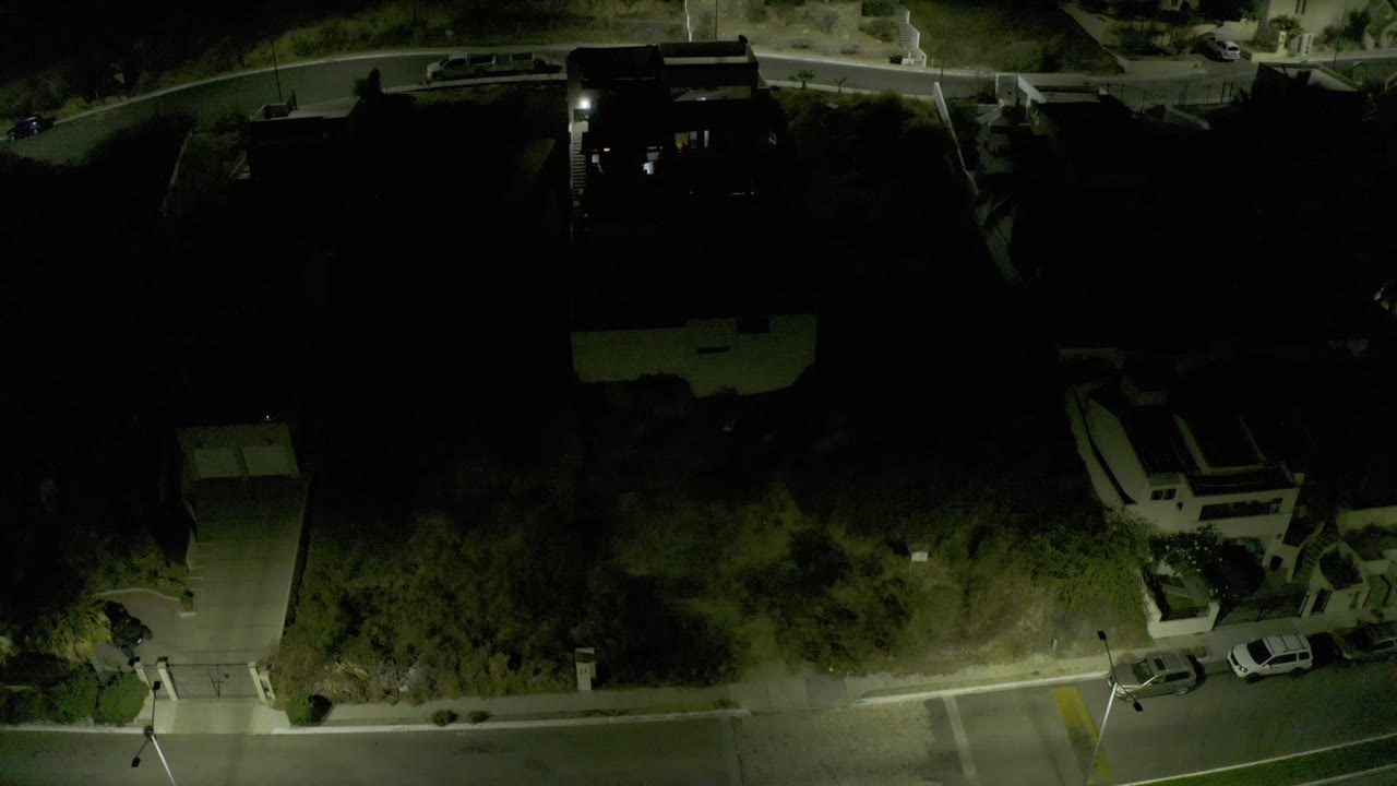 Dim drone view of a residential corner with houses, parked vehicles, and trees.