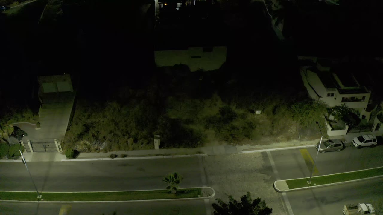 Bird's-eye frame in Los Cabos, Mexico, with two light-colored parked cars near a driveway, dark vegetation above the road, and no visible people.