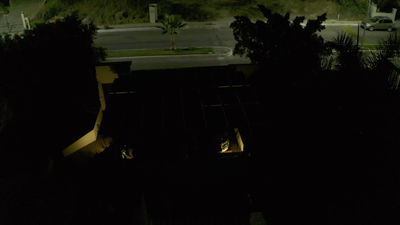 A dark Los Cabos residential exterior seen from above, with a road at the top of frame, dense tree shadows, warm driveway lights, and no visible people.