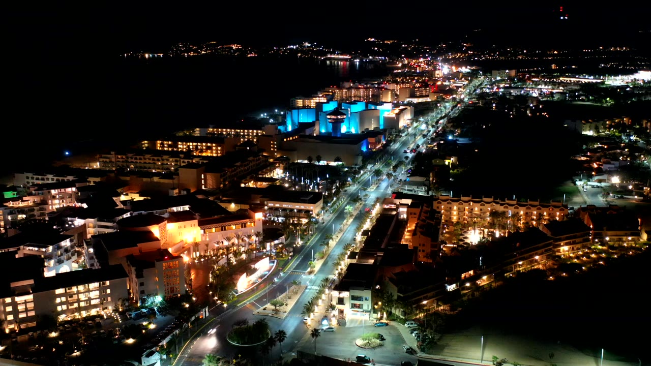Night aerial view over Los Cabos, Mexico, with a bright diagonal road, hotel-like buildings, blue-lit structures, distant shoreline lights, and no visible people.