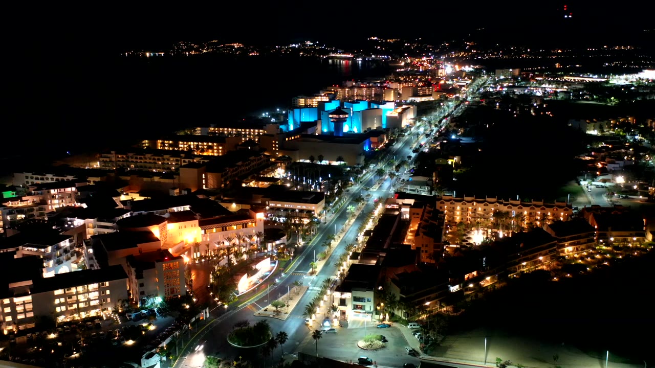 Elevated nighttime frame in Los Cabos with car lights moving along the main road, warm building windows on both sides, blue accent lighting in the midground, and no visible people.