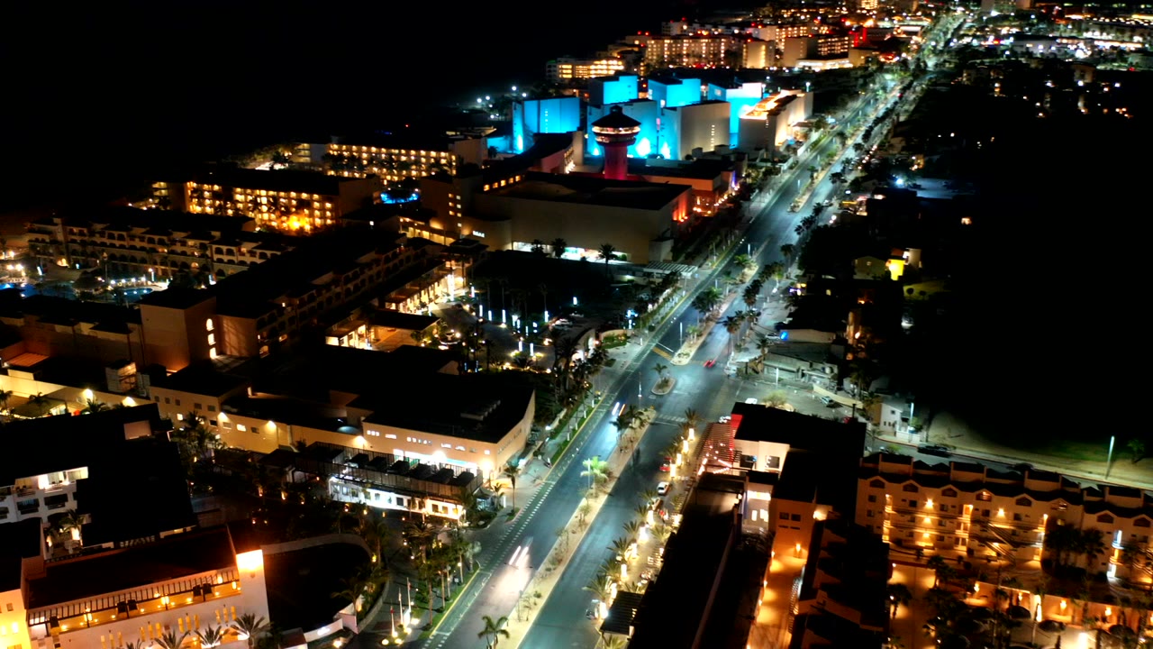 Aerial night view over Los Cabos with a diagonal lit road, warm building lights, blue-lit structures near the top, dark surrounding areas, and no visible people.