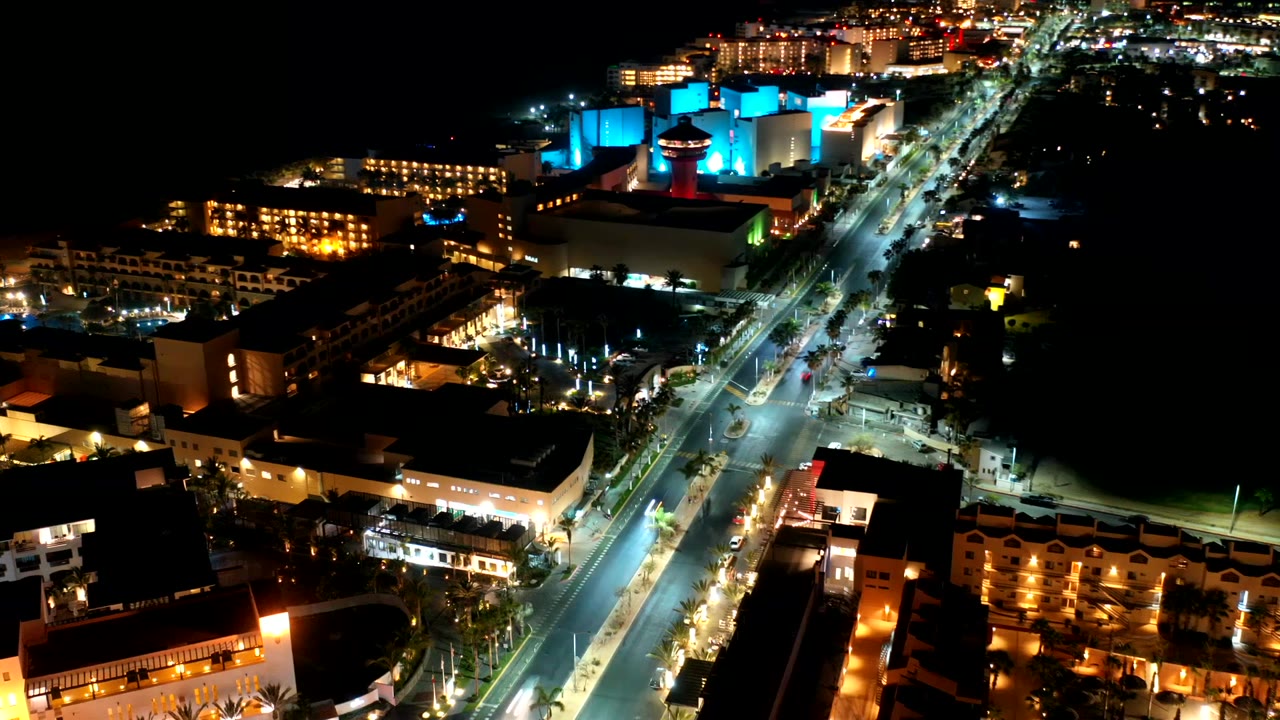 Los Cabos night aerial with traffic lights along the central road, bright intersection areas, warm hotel-like facades, blue accent lighting, and no visible people.