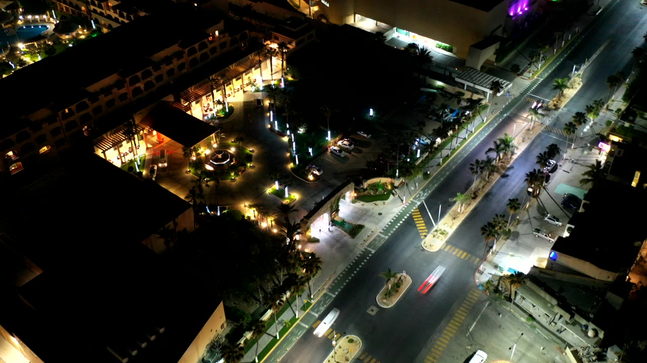 High overhead night view in Los Cabos, Mexico, showing a lit intersection, palm medians, parked cars, and a bright building entrance with no visible people.