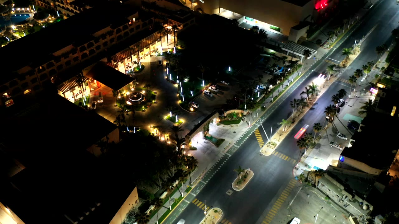 Overhead Los Cabos night frame with the roadway crossing through the center, bright curb lighting, palm-lined medians, parked vehicles, and no visible people.