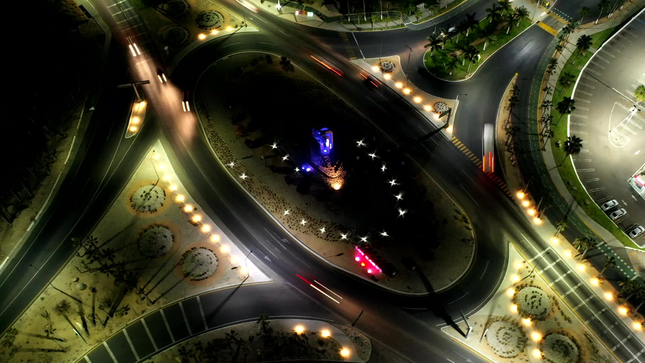 Overhead night frame in Los Cabos, Mexico, showing an oval landscaped traffic island, curving roads, streetlights, vehicle light streaks, and no visible people.
