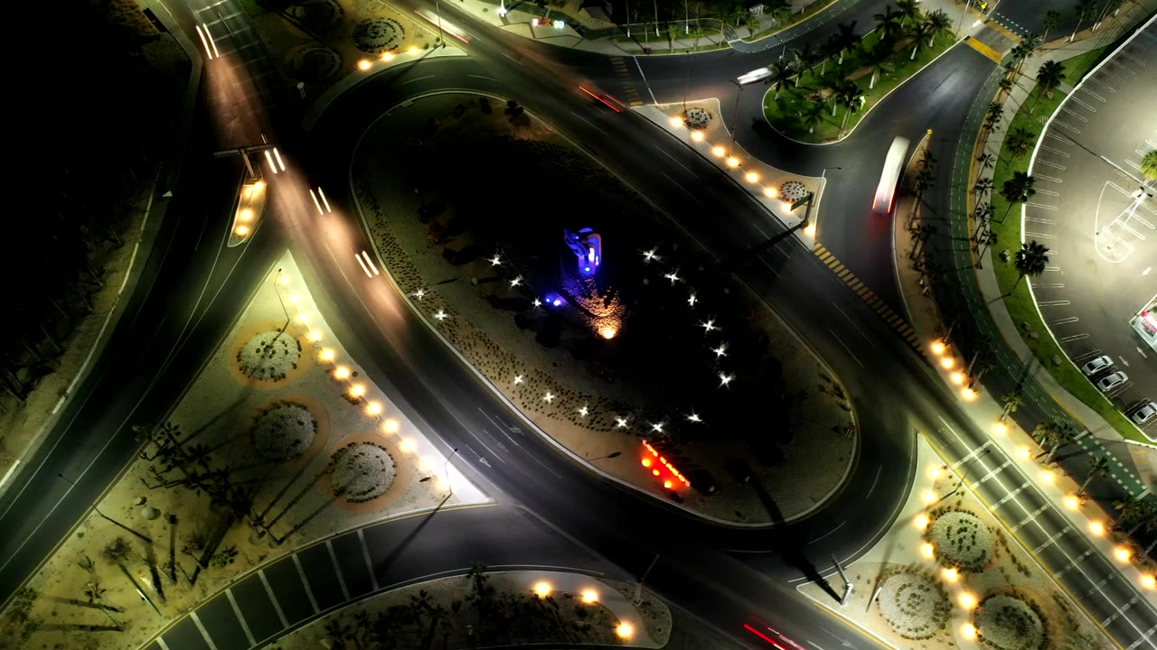 Los Cabos night aerial with the traffic island centered lower in frame, bright streetlamps along the sidewalks, parked cars nearby, moving traffic streaks, and no visible people.