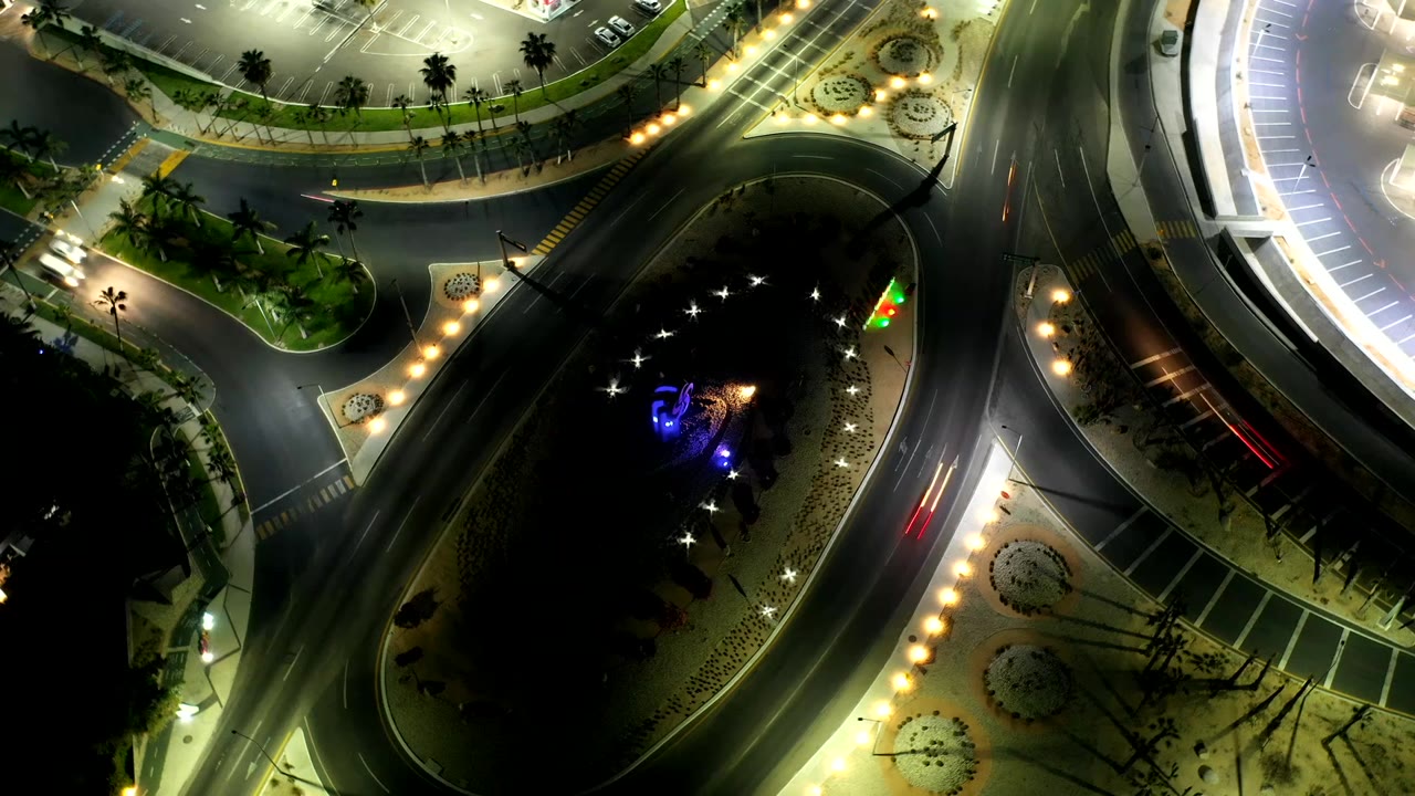 Top-down Los Cabos night view with the oval island lower in frame, bright streetlights along the curb, a parking lot above, and no visible people.