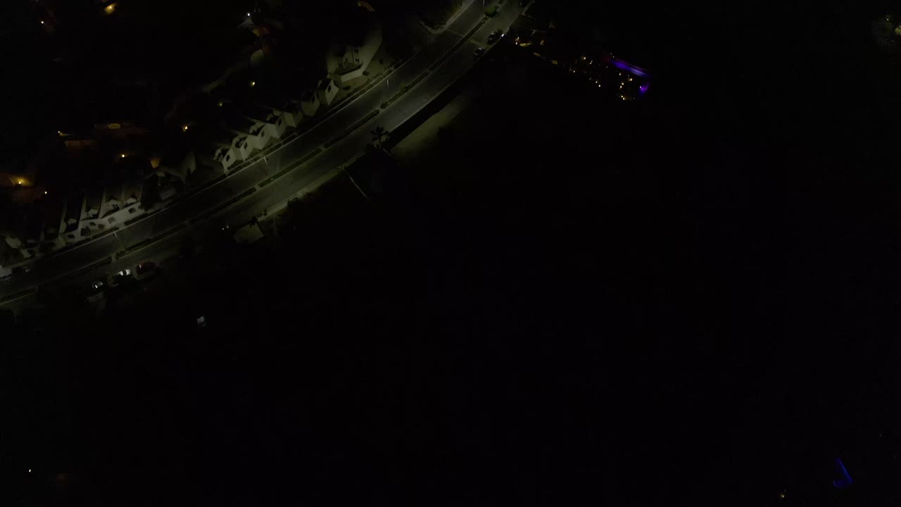 Low-light aerial frame in Los Cabos, Mexico showing a diagonal road, dark houses, a few warm lamps, purple light accents, and no visible people.