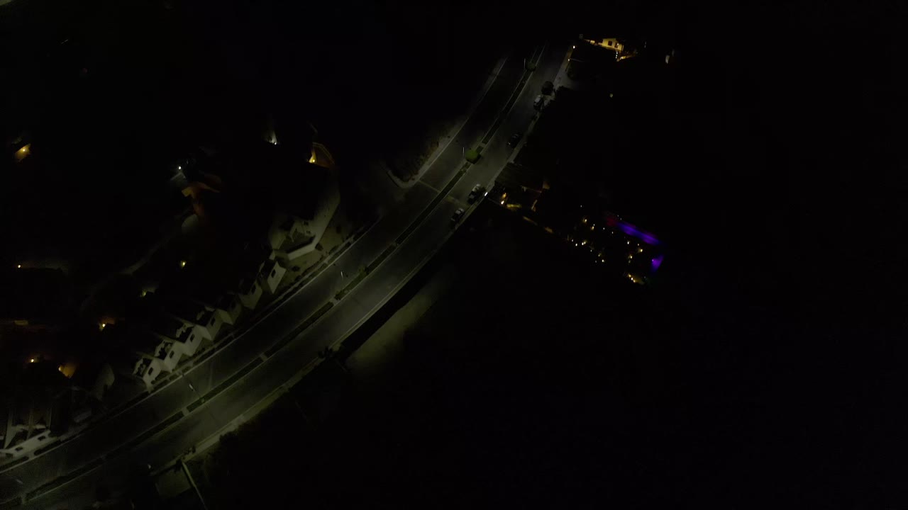 Aerial night view in Los Cabos, Mexico, with the diagonal roadway slightly more centered, faint vehicle lights, nearby structures, and no visible people.