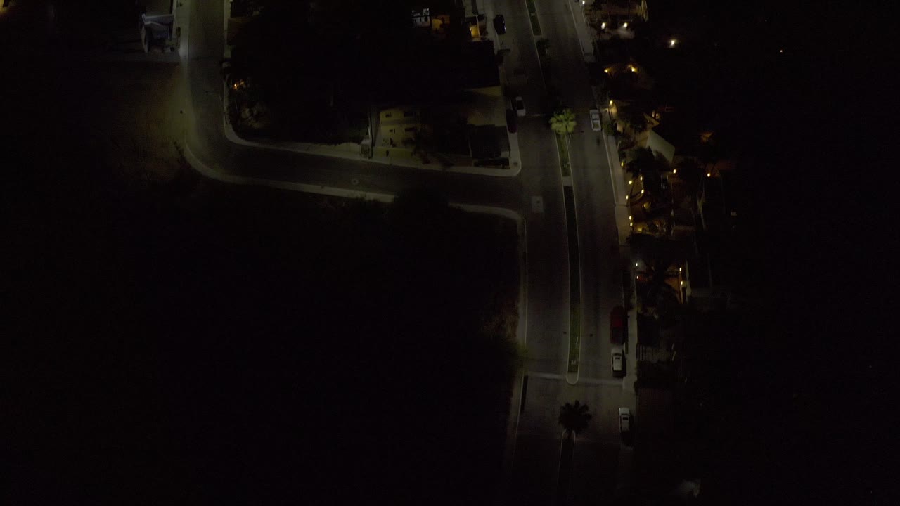 Los Cabos night aerial showing the main road slightly lower in frame, with yellow streetlight pools, dark lots, small buildings, parked vehicles, and no visible people.