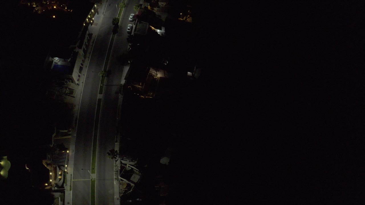 Overhead night view in Los Cabos, Mexico, with a curved road lit by a few streetlights, parked vehicles near buildings, deep surrounding darkness, and no visible people.