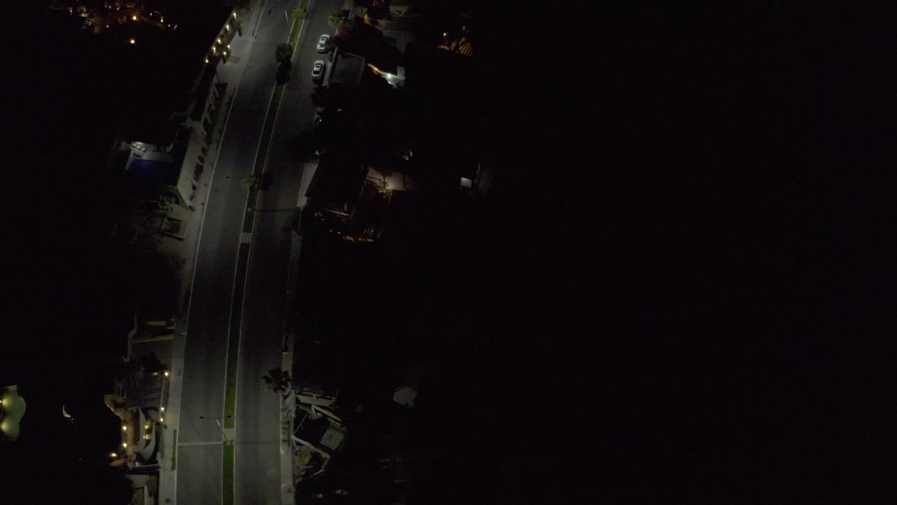 A slightly later overhead frame shows the same Los Cabos road with streetlights falling across the pavement, parked cars near shadowed buildings, and no visible people.