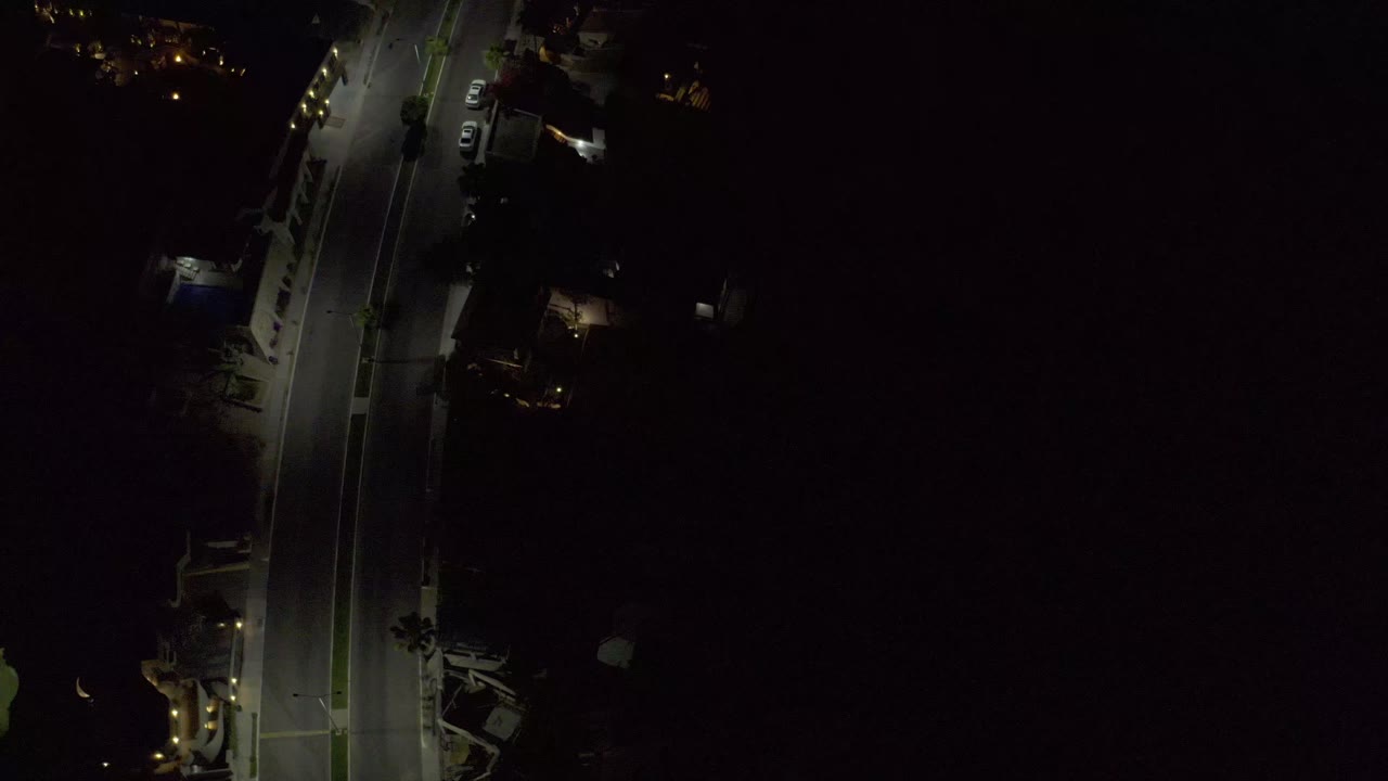 Aerial night view over Los Cabos with the illuminated road still near the left side, small warm lights around buildings, a large dark area, and no visible people.