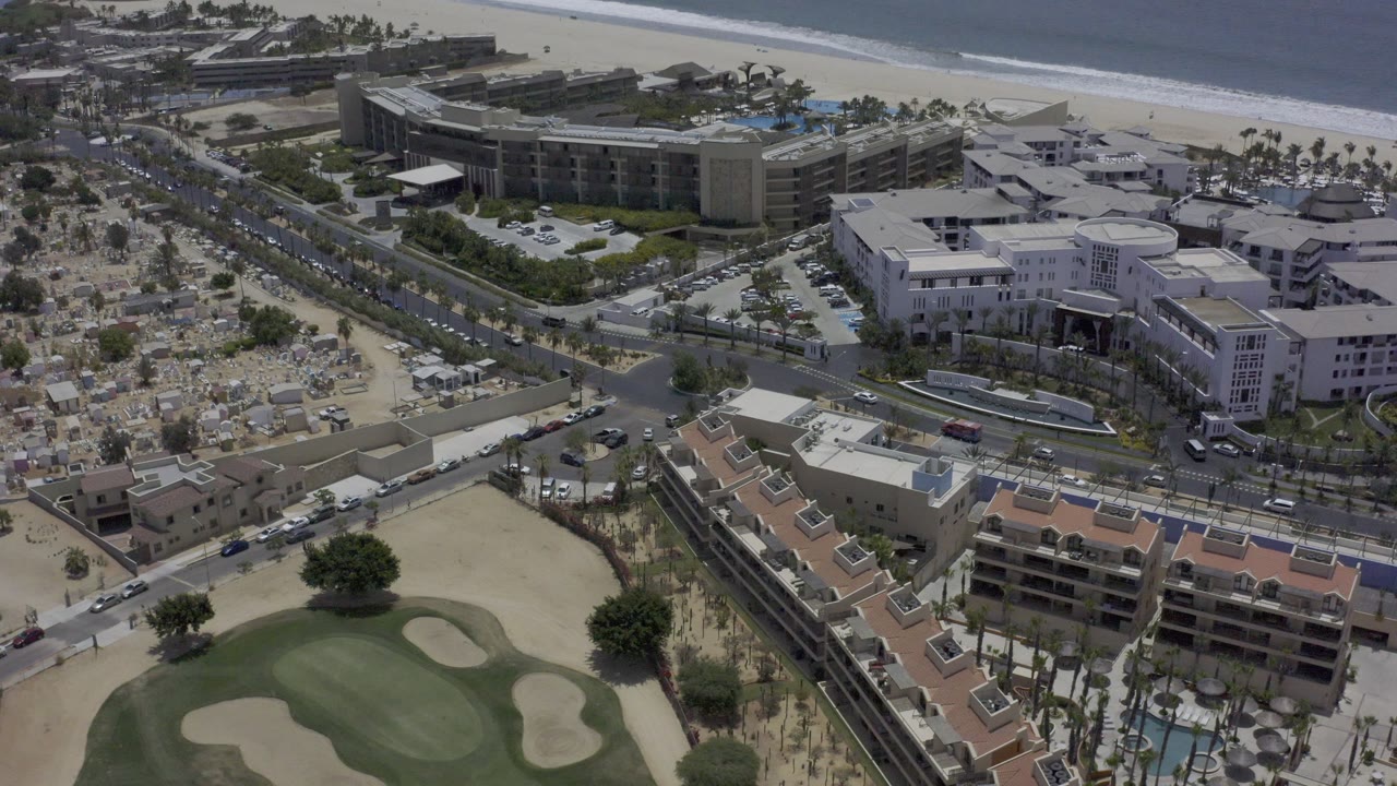 Los Cabos aerial frame showing the golf course lower in the image, a diagonal roadway with small vehicles, clusters of low-rise buildings, the beach, and no visible people.