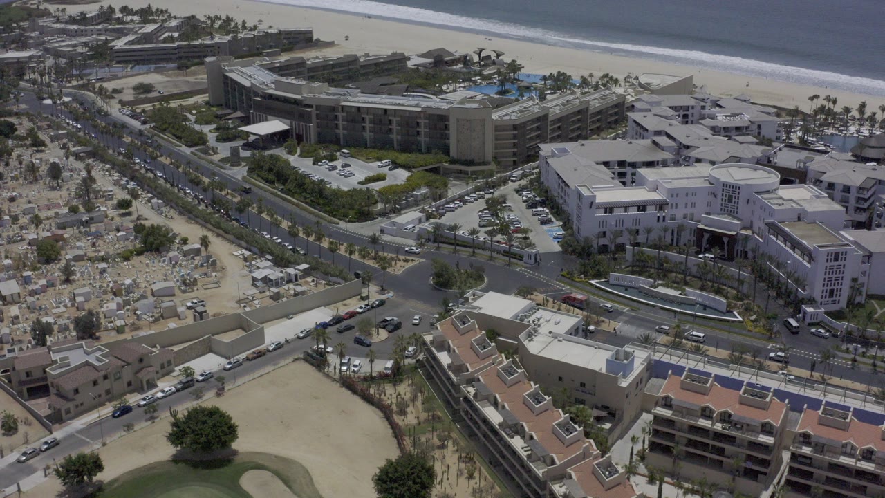 High aerial view over Los Cabos, Mexico, showing beachfront hotels, a road intersection, sandy beach areas, and ocean surf in the background with no visible people.
