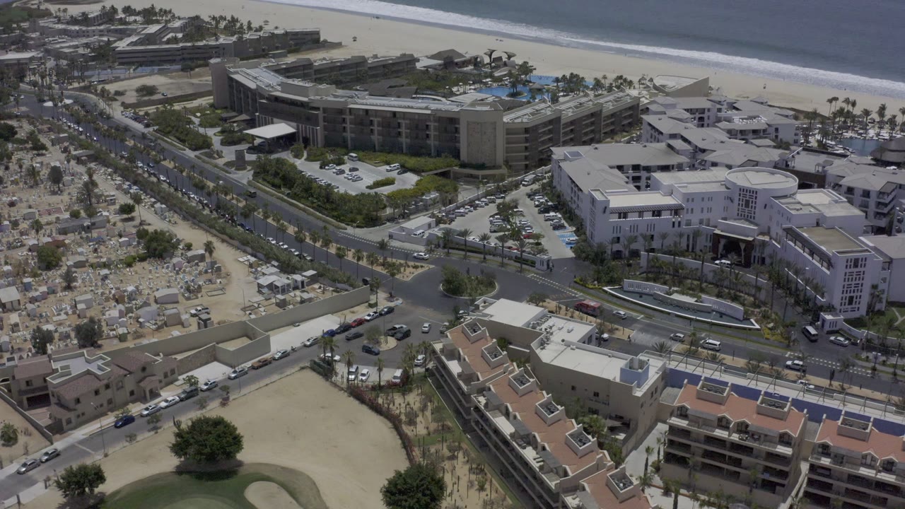 Aerial Los Cabos frame with a large hotel block near the beach, cars along the road, palm-lined grounds, and blue ocean beyond the sand with no visible people.