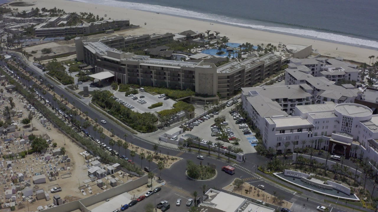Aerial frame in Los Cabos, Mexico showing hotel buildings beside a palm-lined road, sandy beach, ocean surf, and no visible people.