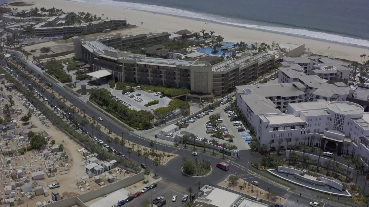 Aerial view over a Los Cabos hotel complex with blue pools, landscaped grounds, parking areas, a wide beach, ocean waves, and no visible people.