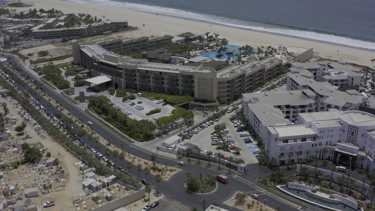 Aerial angle over beachfront hotel, adjacent white buildings, road, beach, and ocean.