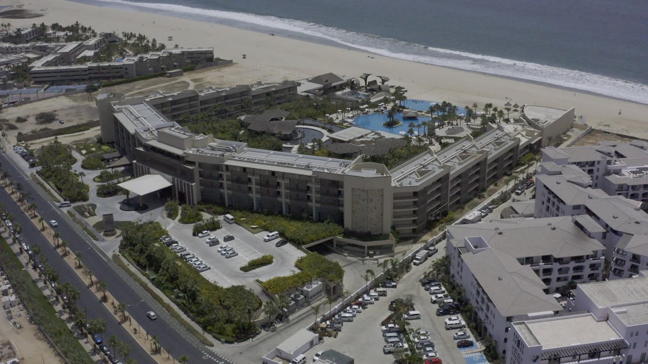 Aerial frame over a Los Cabos beachfront building complex with a blue pool behind the main structure, sandy beach and ocean beyond, road and parking in front, and no visible people.