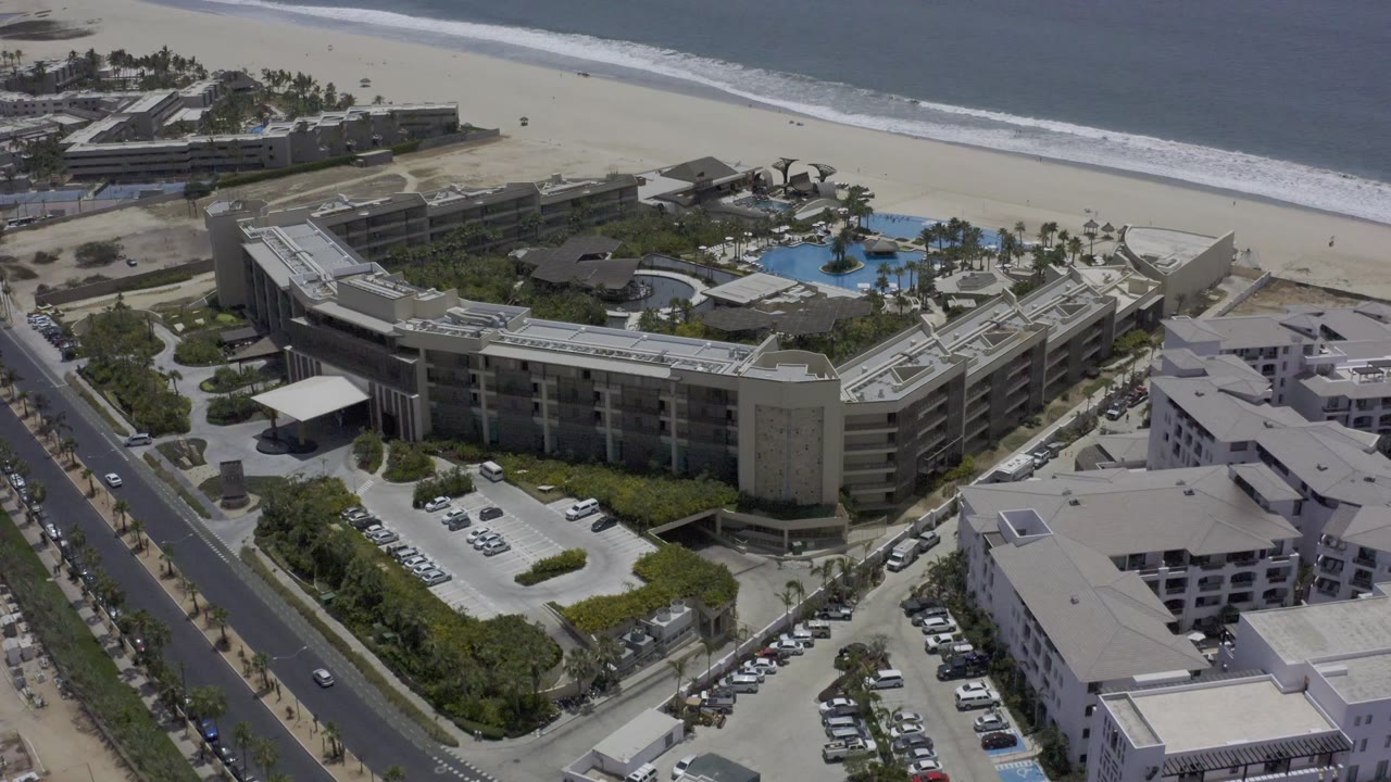 Second aerial frame of the Los Cabos property shows the main building block in the lower frame, the pool courtyard closer to center, ocean surf across the top, and no visible people.