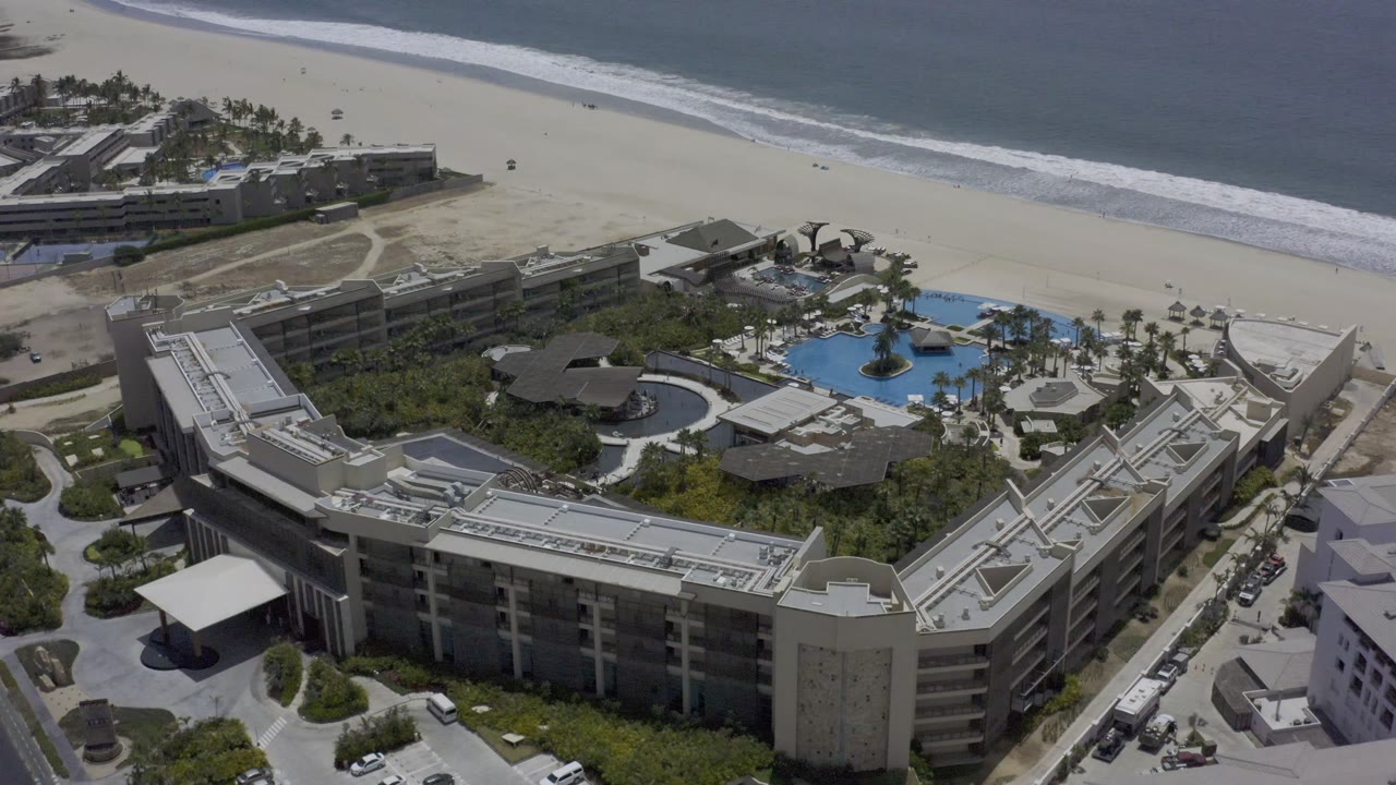 Aerial Los Cabos resort frame at 0:01 with the central pool and palms slightly more centered, hotel roofs in the foreground, beach paths beyond, and small distant people near the shoreline.