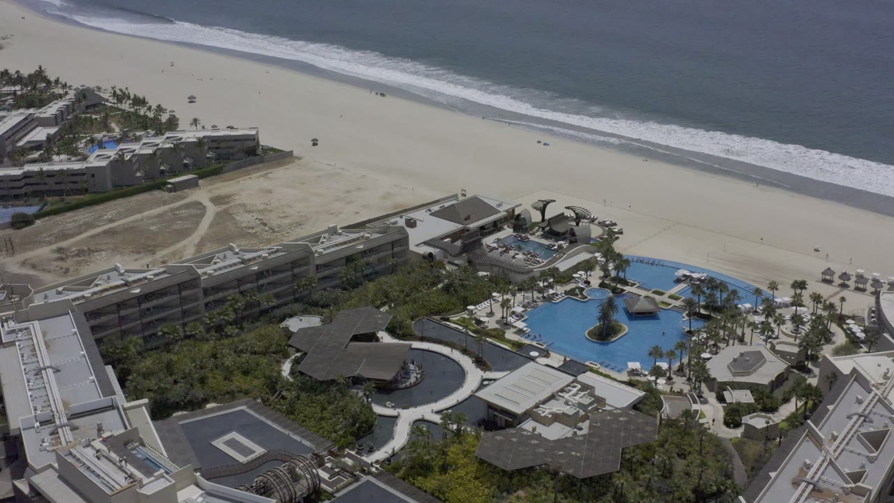 High coastal view with resort buildings, blue pools, beach, and surf.