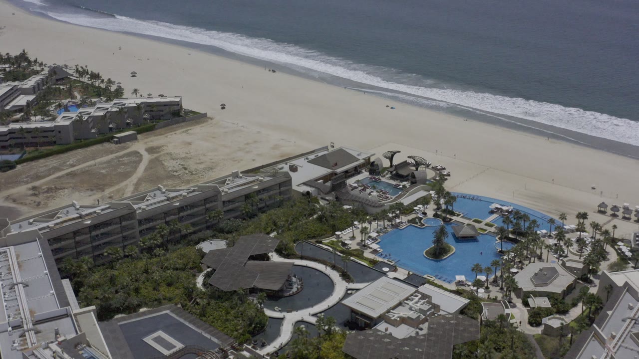 Drone-like Los Cabos view with angular resort buildings, a curved pool area, palm rows, open sand, and darker blue ocean at the top of frame.