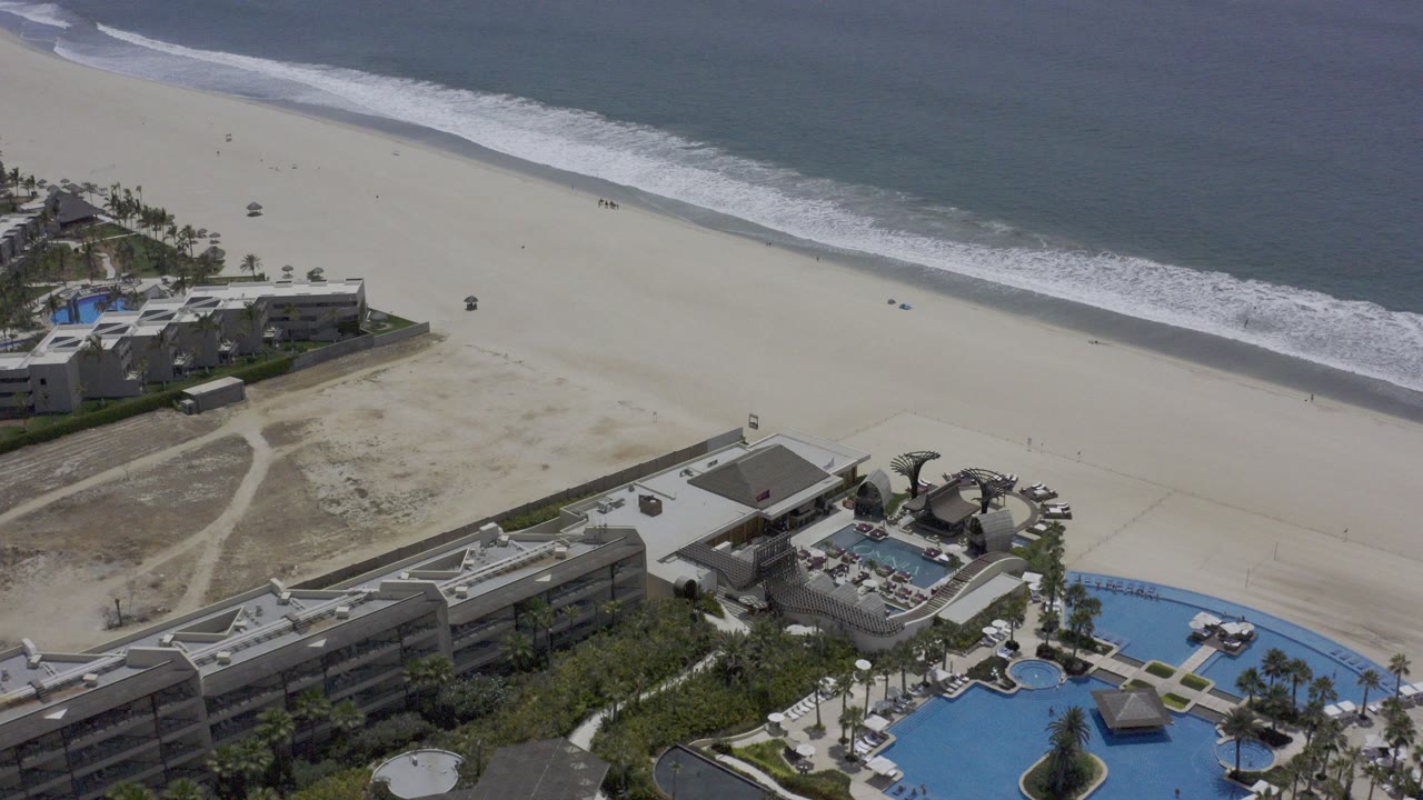 Aerial frame over a Los Cabos beach with pale sand, a diagonal surf line, resort buildings and pools at the bottom edge, and no clearly visible people.