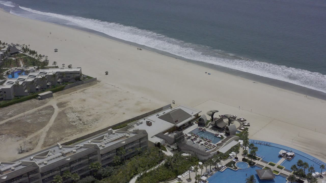 Aerial shoreline view with beachfront architecture, pools, sand, and ocean.