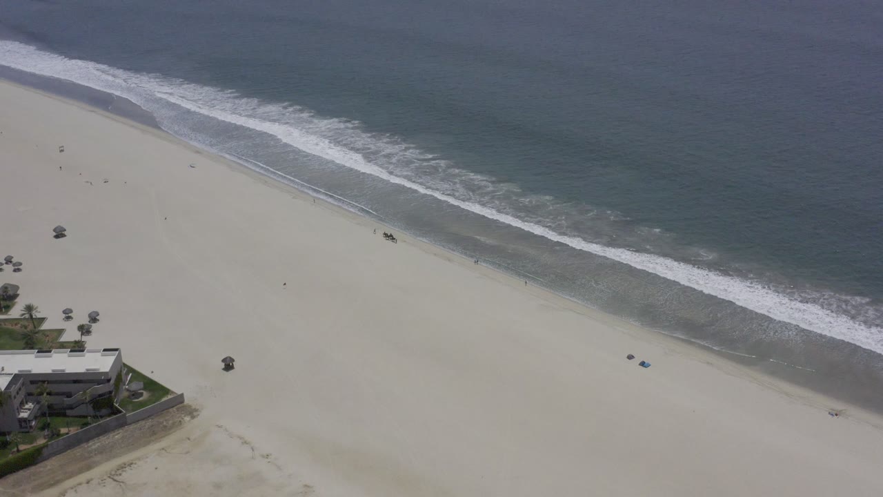 Overhead Los Cabos beach frame with the foamy wave band slightly lower across the shoreline and a small beachfront structure near the bottom edge.