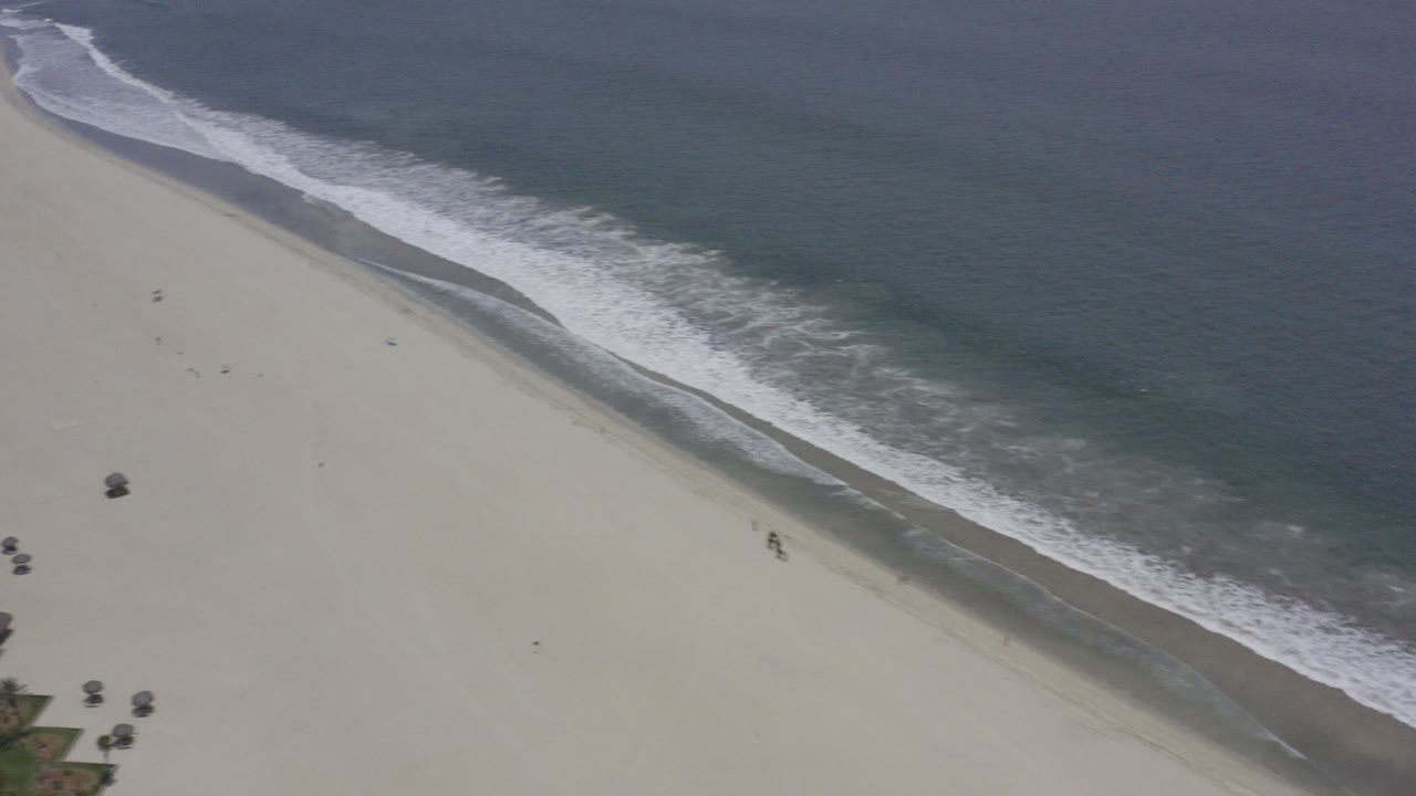Aerial view over Los Cabos showing a wide pale beach, a diagonal white surf band, and dark blue-green sea with no recognizable people.
