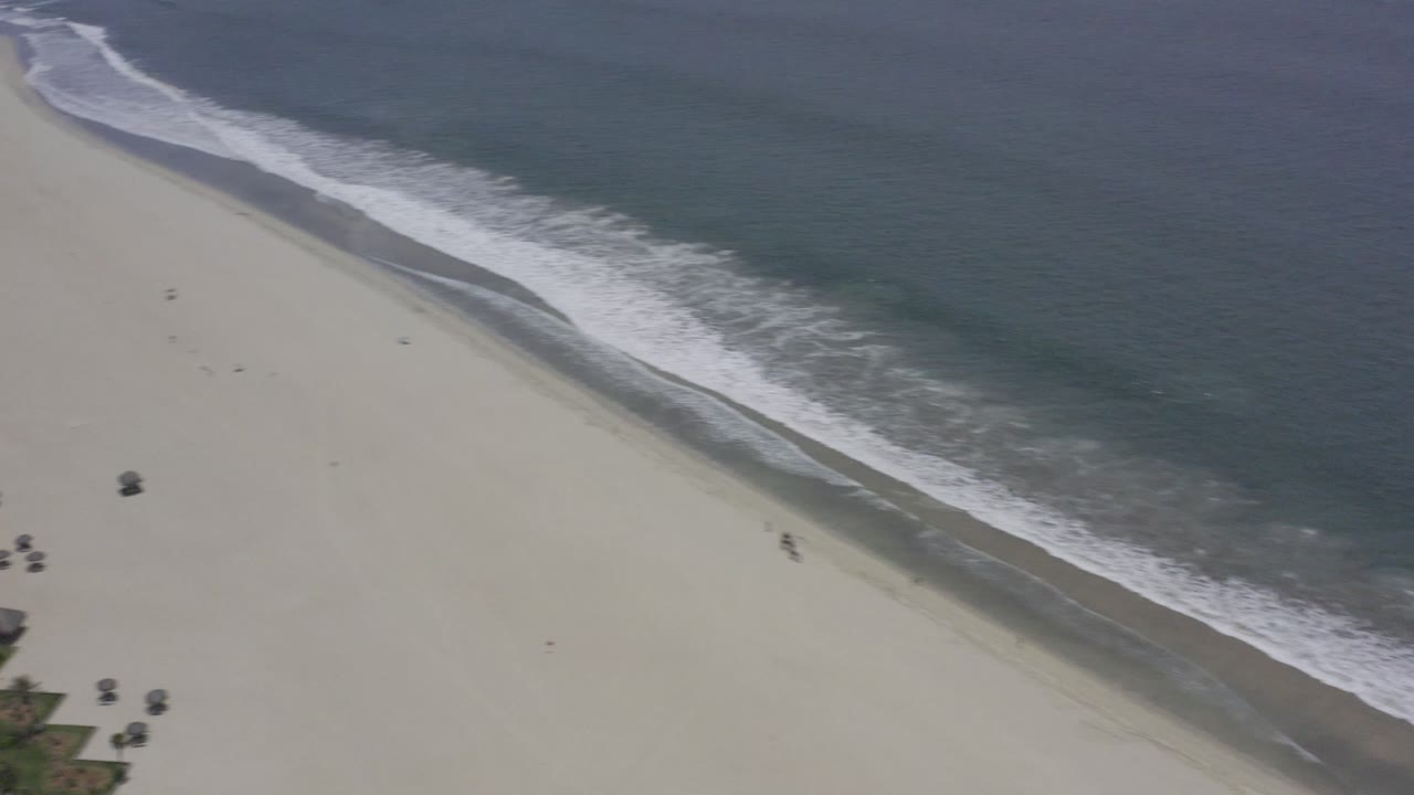 Aerial Los Cabos shoreline frame with white waves tracing the sand and a few tiny distant objects on the open beach.