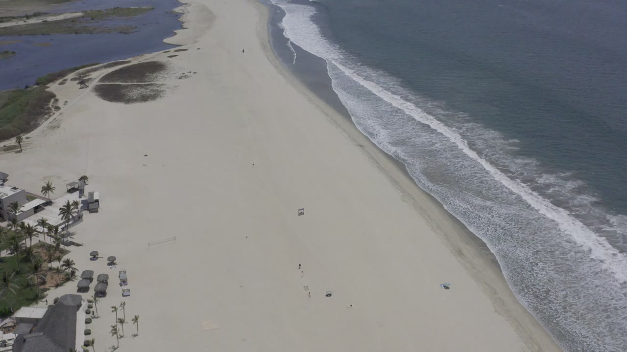 Aerial Los Cabos coastline view with pale sand dominating the frame, foamy waves tracking the right edge, dark vegetation patches inland, and no visible people.