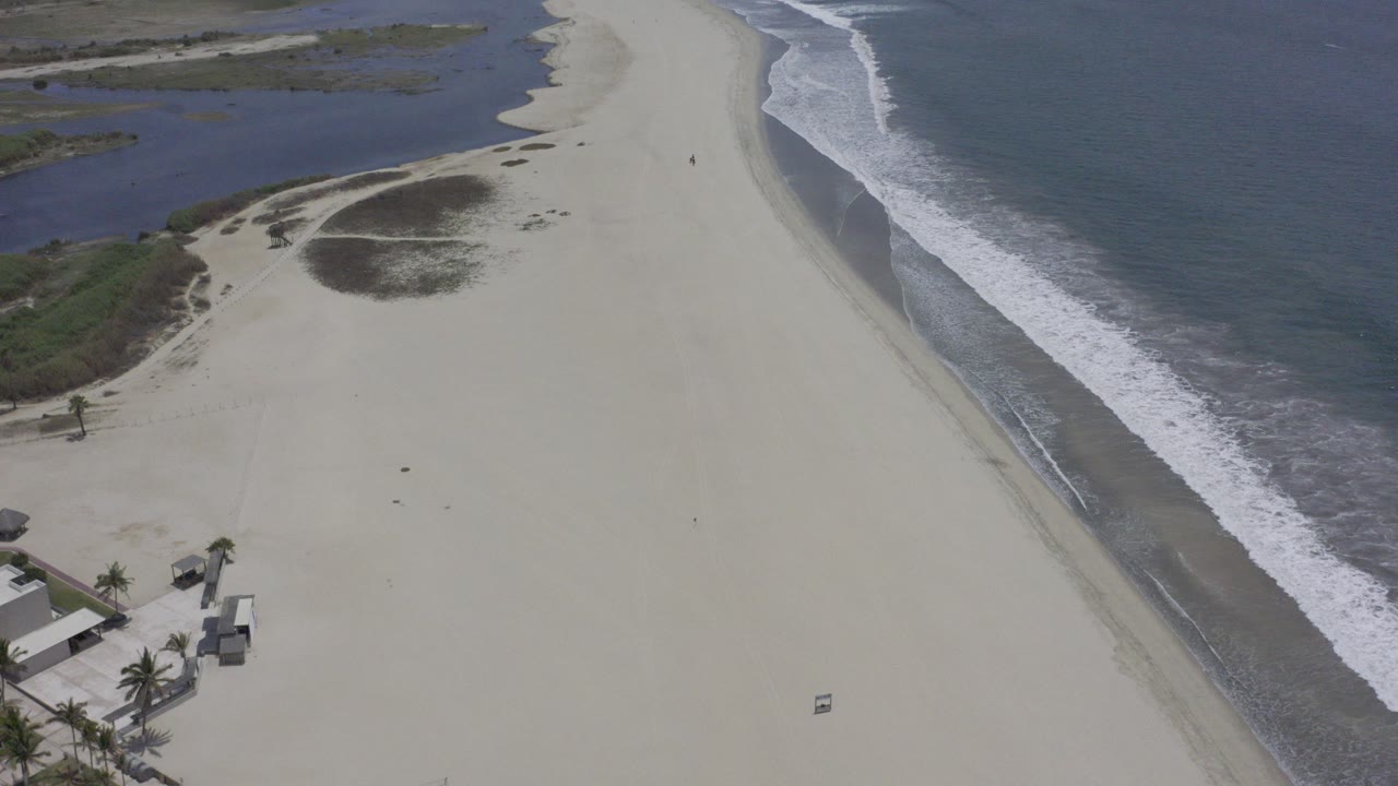 Aerial view over a wide Los Cabos beach with white surf on the right, dune vegetation and an inlet behind the sand, small structures near the lower edge, and no visible people.