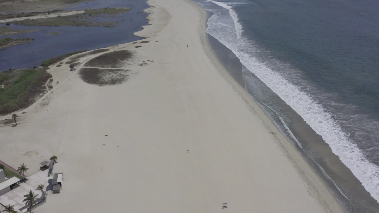 Aerial beach view with ocean surf and buildings at the lower shoreline.