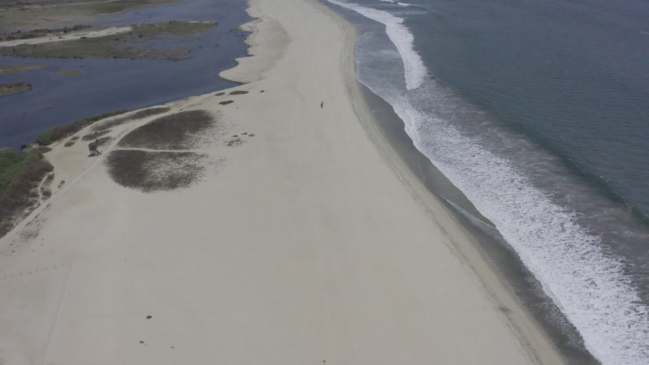 High aerial view in Los Cabos, Mexico, showing a pale sand strip between darker water on the left and breaking ocean surf on the right, with no visible people.