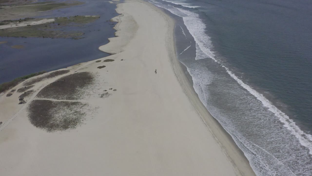 Los Cabos shoreline seen from above, with white surf lines beside a wide pale beach and lagoon water at the upper left, no visible people.